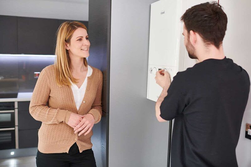 An engineer adjusting a wall-mounted Ideal combi boiler while a woman watches in a modern kitchen