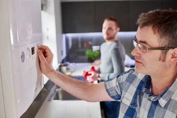 A man adjusting the controls of a wall-mounted combi boiler in a kitchen while another man stands behind him
