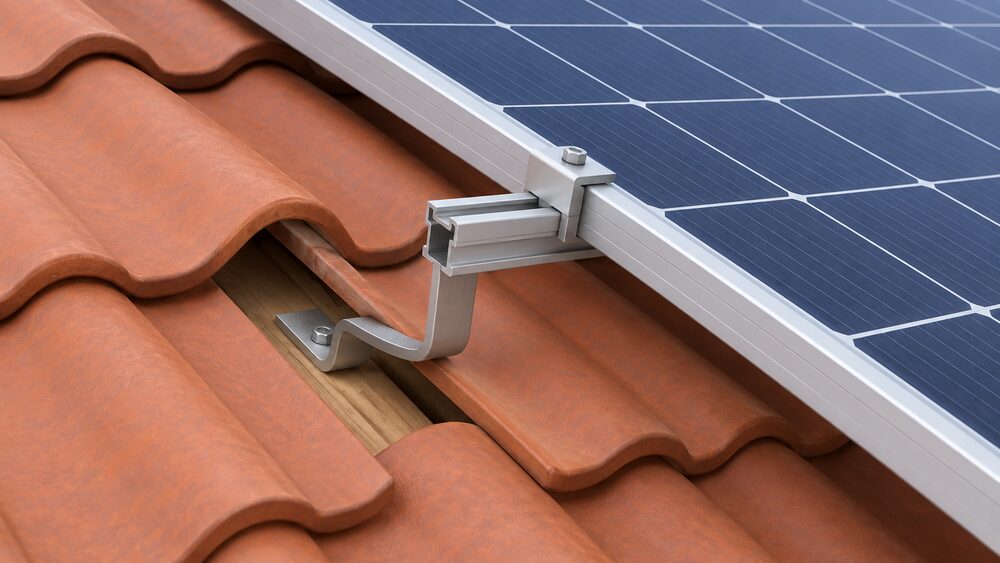 A close-up of a metal roof hook fixed to a rafter under a lifted pan tile on a UK tiled roof, bridging out over the tile with an aluminium mounting rail bolted along its top, with a middle clamp gripping a solar module frame above the tile covering.