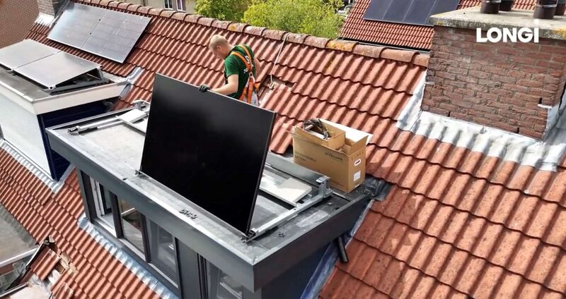 An installer on a tiled Dutch roof lifting a LONGi solar panel into place on a flat-roof mounting frame, with other panels already installed