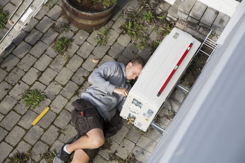 An installer lying on a block-paved patio working on an outdoor heat pump unit beside a house wall