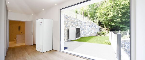 An air source heat pump outdoor unit mounted on a stone wall outside a modern home, with an indoor unit visible in the hallway