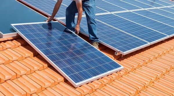 A worker positioning a solar photovoltaic panel on a terracotta-tiled roof alongside already installed panels