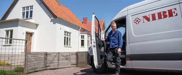 A NIBE installer in blue workwear stepping out of a branded white van outside a white house with a red tiled roof