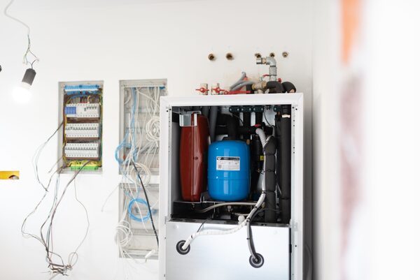 An open heat pump indoor unit with expansion vessel and pipework being installed on a wall next to exposed electrical wiring