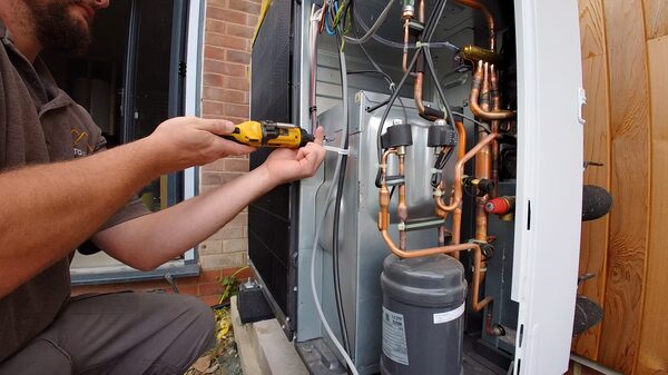 An engineer using a screwdriver to service the exposed pipework and components of an air source heat pump unit outside a house