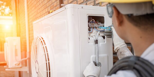 An engineer in a hard hat and gloves wiring an outdoor air source heat pump unit against a brick wall