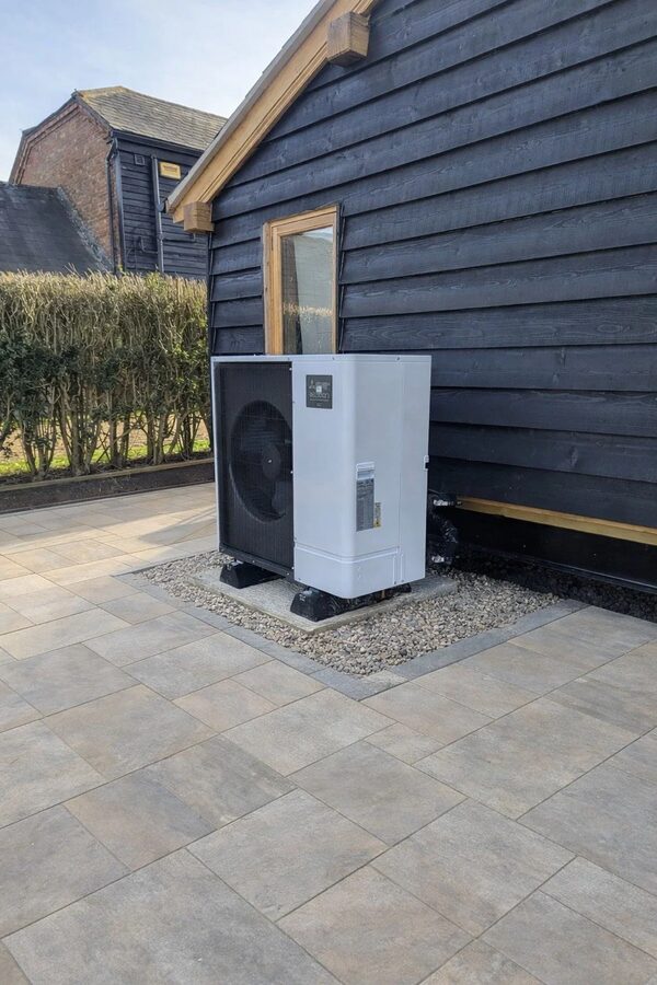 An air source heat pump unit installed on a gravel base outside a black timber-clad house