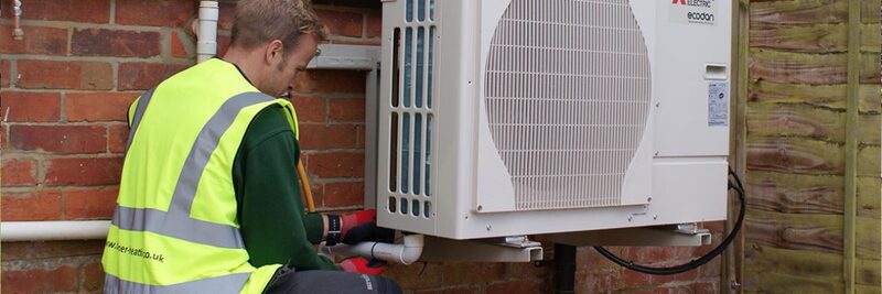 An engineer in a high-visibility vest working on a Mitsubishi Ecodan air source heat pump mounted on a brick wall outside a house