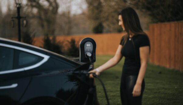 A woman plugging a charging cable into a black electric car beside a wall-mounted myenergi zappi EV charger in a garden driveway