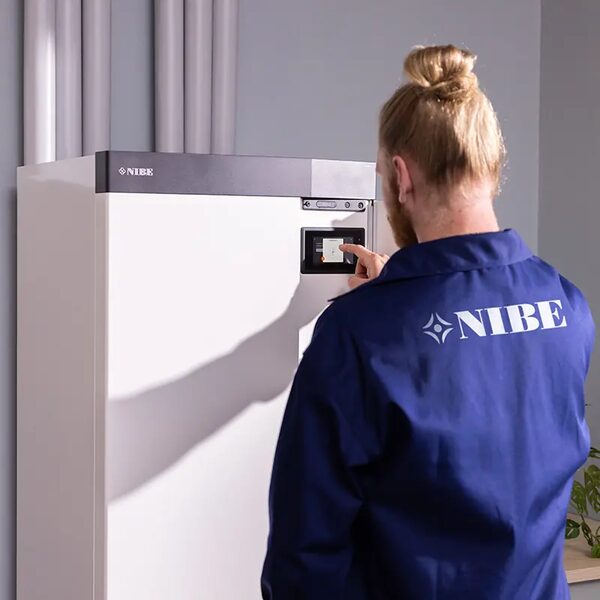 A technician in a NIBE-branded jacket adjusting the control panel of an indoor NIBE heat pump unit