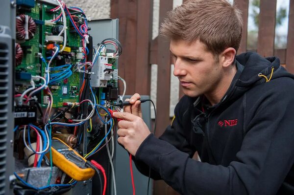 A technician in a black NIBE hoodie using a multimeter to test the wiring inside a heat pump unit outdoors