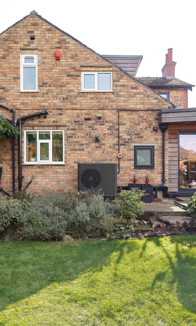 A dark grey Aira air source heat pump unit installed on the exterior brick wall of a house beside a lawn