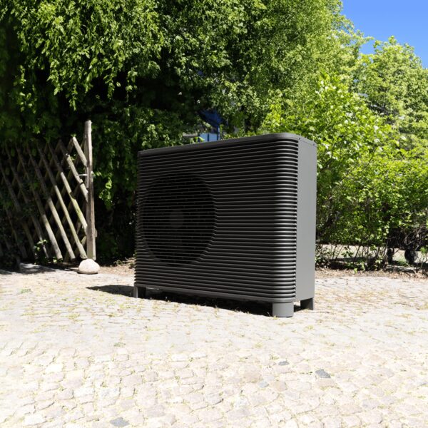 A dark grey Aira air source heat pump outdoor unit standing on cobbled paving in a garden with trees and a wooden trellis behind