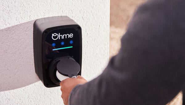 A person plugging a charging cable into a black Ohme wall-mounted EV charger on a white exterior wall