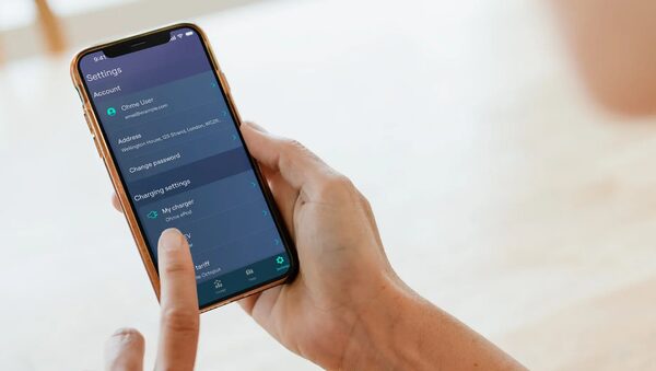 A hand holding a smartphone showing the Ohme EV charging app settings screen