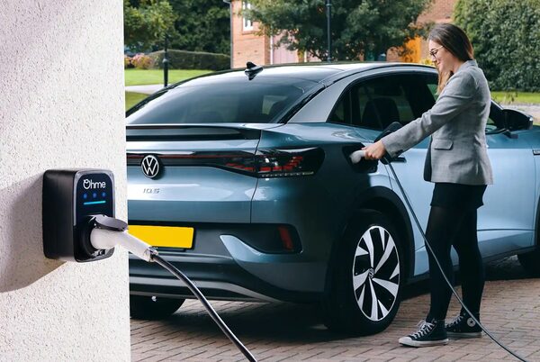A woman plugging a charging cable into a blue Volkswagen ID.5 electric car on a driveway, next to a wall-mounted black Ohme home EV charger