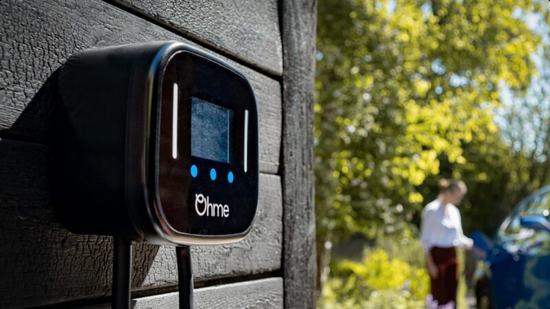 An Ohme home EV charger mounted on a dark wooden wall with a woman and car blurred in the background