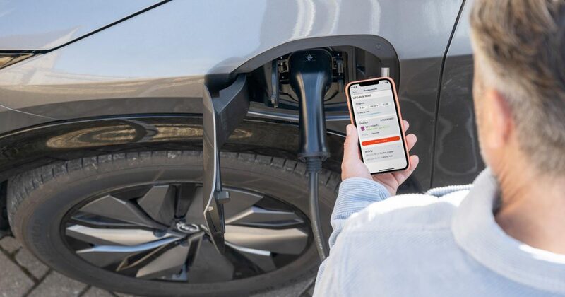 A person holding a smartphone showing a charging app next to an electric car plugged in with a charging cable