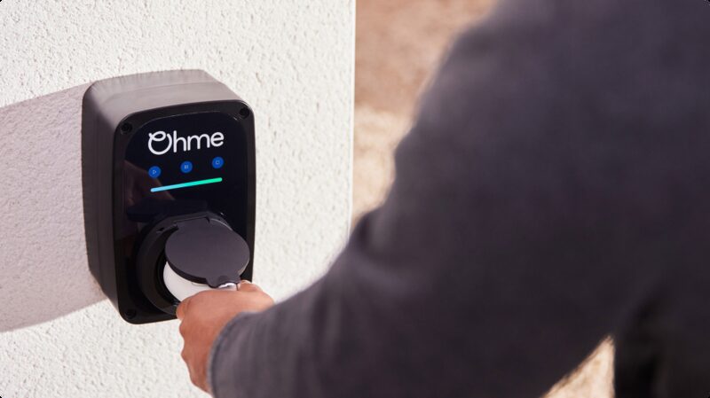 A person plugging a charging cable into a black Ohme ePod EV charger mounted on a white wall