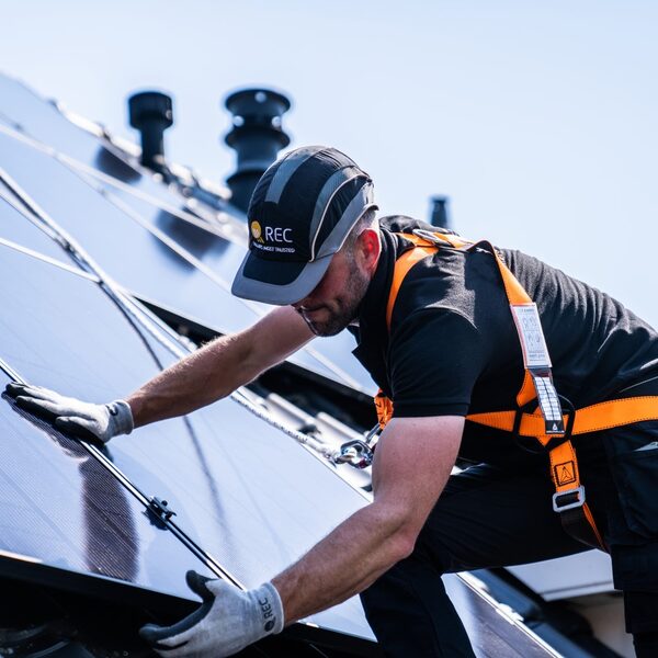 An installer in a REC-branded cap and orange safety harness fitting solar panels on a roof