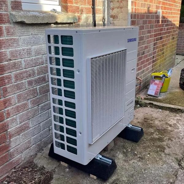 A white Samsung air source heat pump outdoor unit installed on mounts against a brick house wall