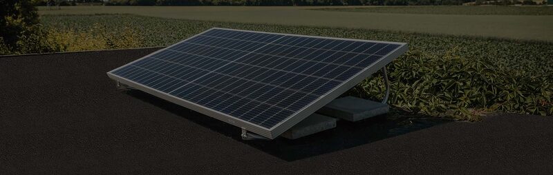 A solar panel mounted on a flat-roof ballasted mounting ramp on a dark flat roof with fields behind