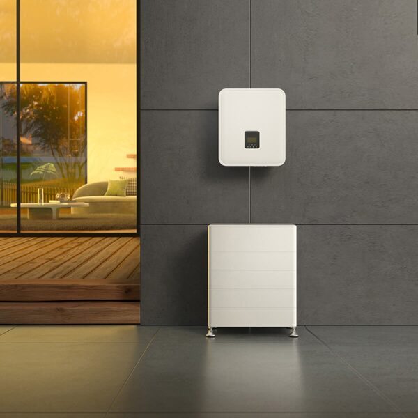 A white wall-mounted hybrid solar inverter above a stacked floor-standing home battery on a grey wall beside a modern living room