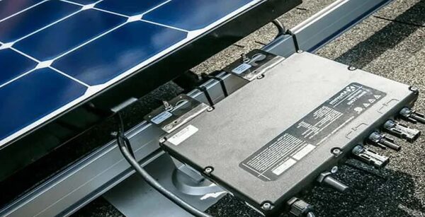 A micro inverter mounted on a rail beneath a solar panel on a shingled roof