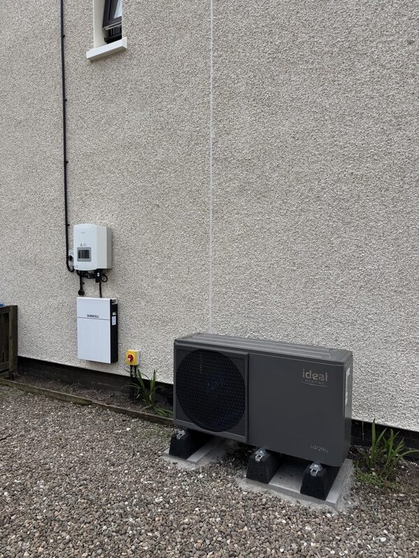 An Ideal air source heat pump unit on the ground outside a house with a Sunamp thermal battery and inverter mounted on the wall above