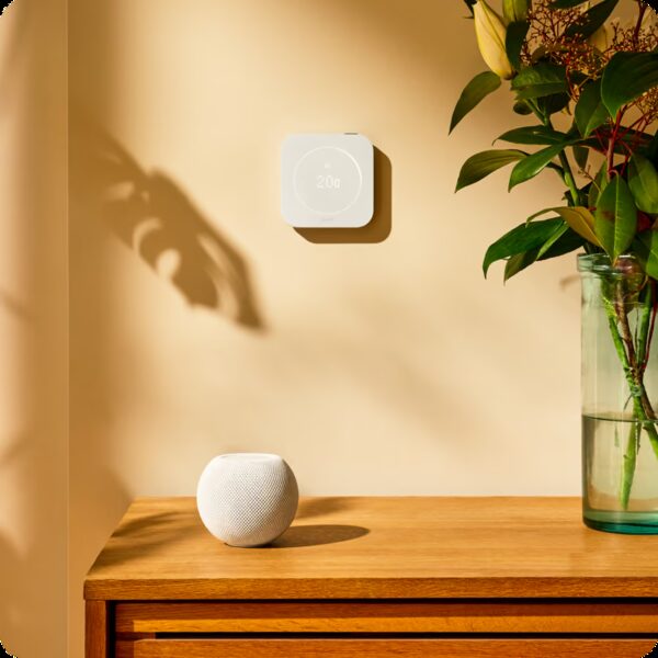 A white tado smart thermostat mounted on a beige wall showing 20 degrees, above a wooden sideboard with a HomePod mini smart speaker and a vase of flowers