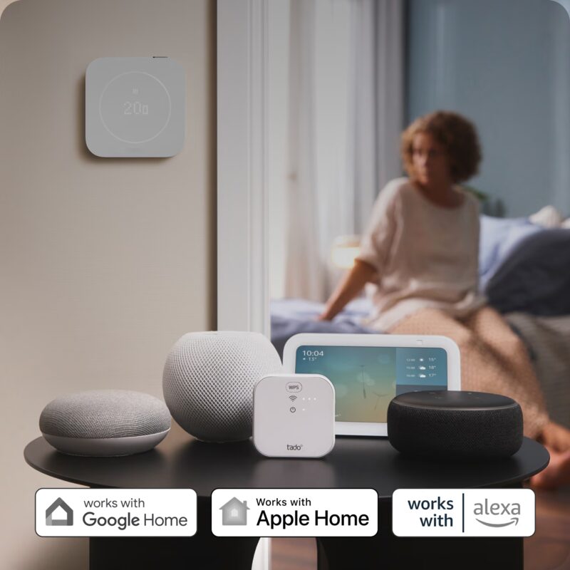 A tado smart thermostat on a wall and a tado bridge on a table beside smart speakers and a display, with 'works with Google Home, Apple Home and Alexa' badges, in a bedroom