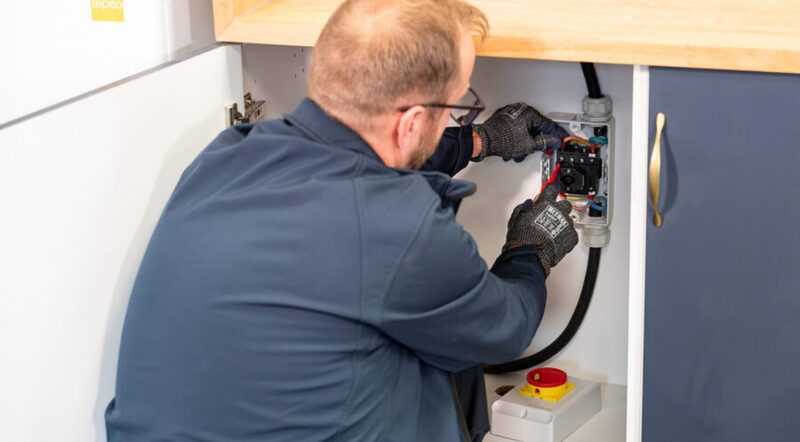 A technician in a navy uniform servicing electrical wiring inside a kitchen cupboard, likely a ZEB boiler installation