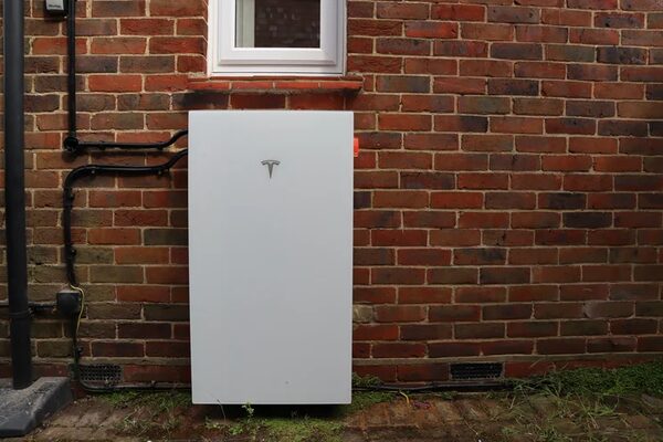 A white Tesla Powerwall home battery mounted on the exterior brick wall of a house