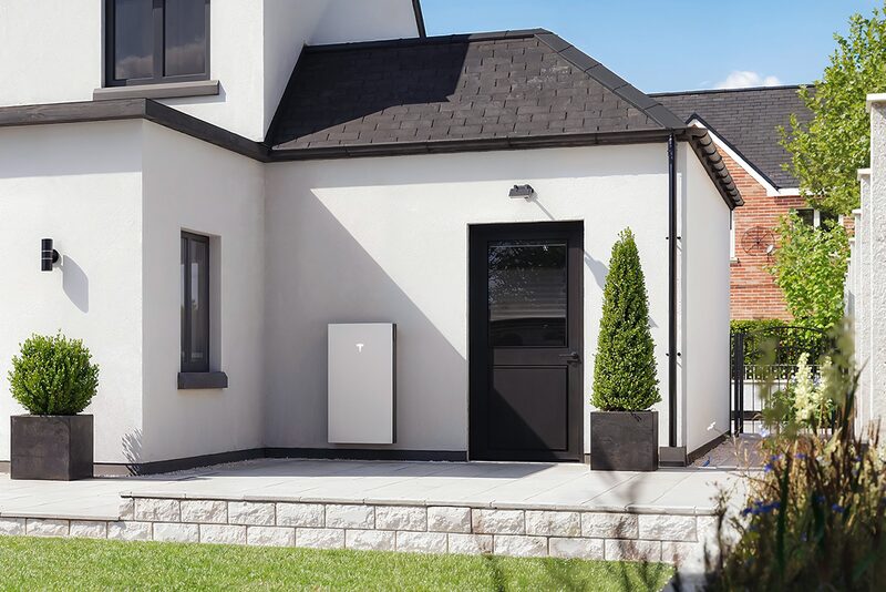 A white Tesla Powerwall battery mounted on the exterior wall of a modern house beside a black door