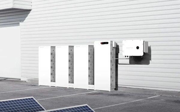A row of white Sigenergy home battery stack units installed against an exterior wall next to a wall-mounted inverter, with solar panels in the foreground