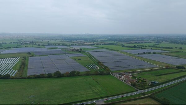 An aerial view of a large solar farm with rows of ground-mounted solar panels spread across green fields beside a road