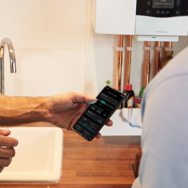A person holding a smartphone showing a heating control app in front of a wall-mounted Vaillant ecoTEC plus boiler in a kitchen