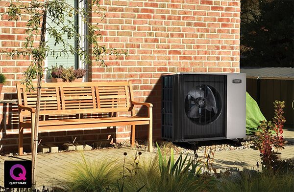 An air source heat pump installed outside a brick house next to a wooden garden bench, with a Quiet Mark logo in the corner