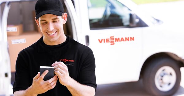 A smiling Viessmann service engineer in branded black uniform using a smartphone in front of a white Viessmann van