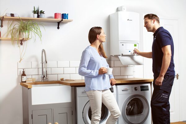 A Worcester Bosch engineer in branded uniform explaining a wall-mounted combi boiler to a woman in a utility room with washing machines