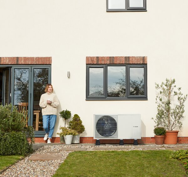 A Worcester Bosch air source heat pump installed on the outside wall of a house, with a woman standing nearby holding a mug