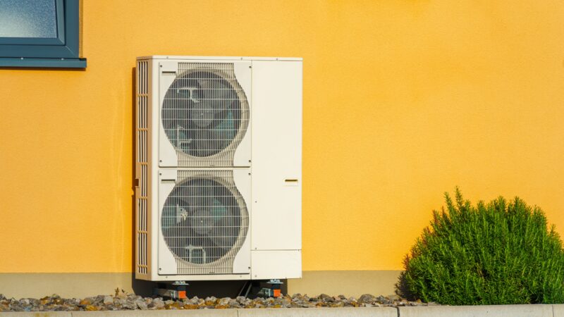 An air source heat pump unit mounted on the outside wall of a yellow house next to a green shrub