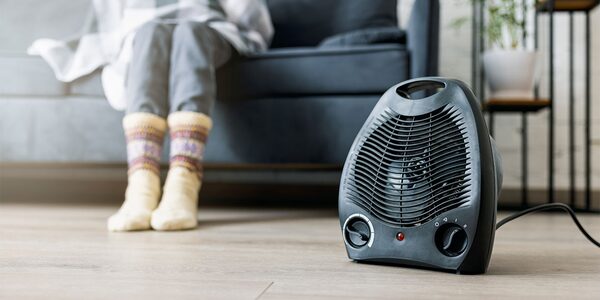 A black portable electric fan heater on a wooden floor with a person in cosy socks resting their feet on a sofa behind it