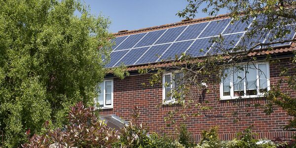 Solar panels installed on the tiled roof of a brick house surrounded by trees and garden plants