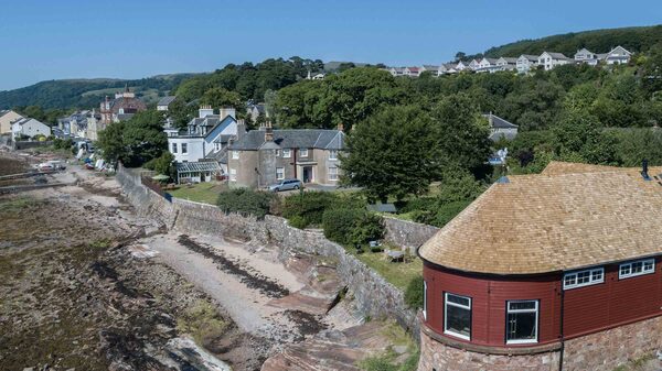 A red-painted coastal house with a cedar shingle roof overlooking a rocky shore, with other houses and hills behind