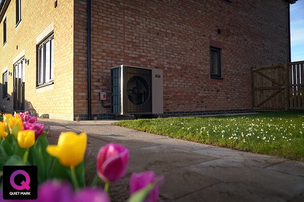 An air source heat pump unit installed on the exterior brick wall of a house, with a garden path and tulips in the foreground