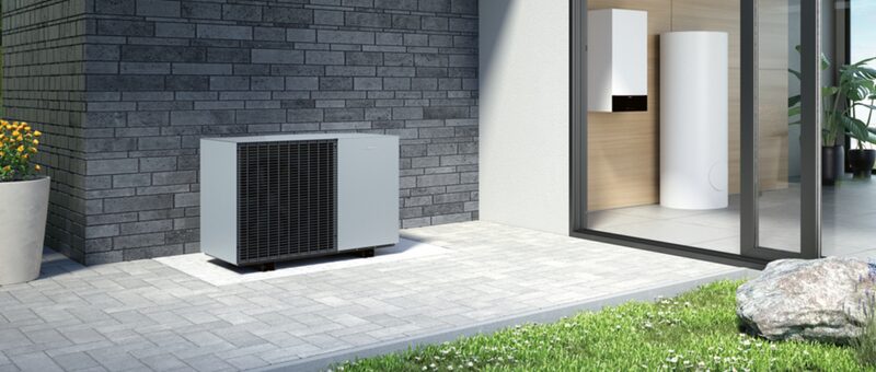 An air source heat pump unit installed outside a modern grey brick house, with a hot water cylinder and wall-mounted unit visible inside through the glass doors