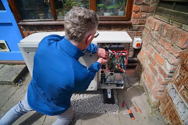 A technician working on the open wiring of an air source heat pump unit outside a brick house