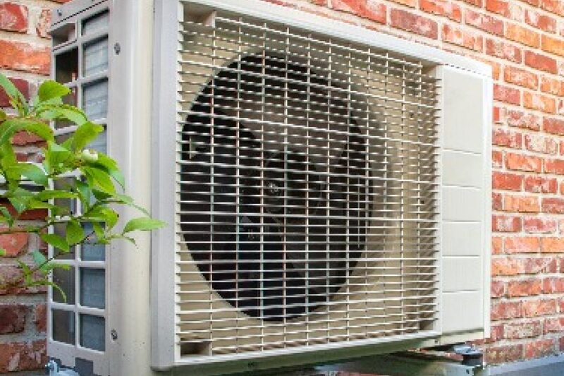 An air source heat pump unit mounted on the outside brick wall of a house, with a plant in the foreground